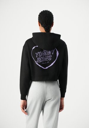 Melnā īsā kapučjakā ar purpura sirds grafiku, uz kuras ir uzraksts "TOMMY JEANS Kopš 85", aizmugurē, saskaņota ar pelēkām sporta biksēm.