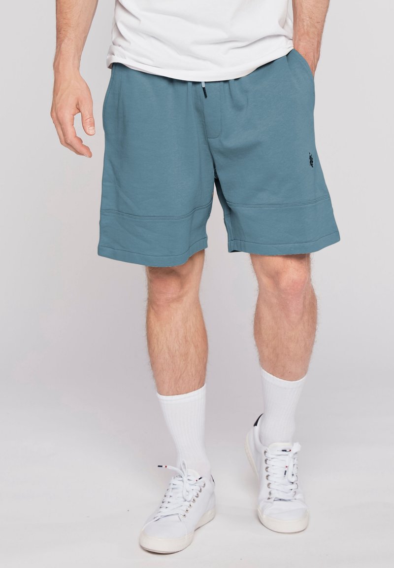 Homme portant un short décontracté bleu, des chaussettes blanches montantes, des baskets blanches et un t-shirt blanc, debout avec les mains près des poches sur un fond uni.