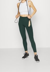 Vit atletisk croptopp med blå ränder, gröna högmidjade leggings och svarta sneakers, håller en hoppselekabel med trähandtag.