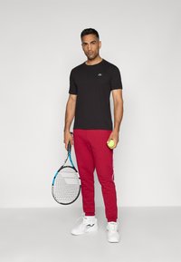 Black short-sleeve T-shirt, red sweatpants with white side stripes, white athletic shoes, holding a tennis racket and a green ball.