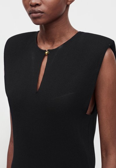Femme portant une robe noire texturée sans manches avec une petite fente à l’avant, fermée par un seul bouton doré au niveau de l’encolure.