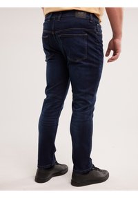 Bărbat purtând blugi skinny albastru închis și adidași negri, văzut din spate pe un fundal simplu, deschis la culoare.