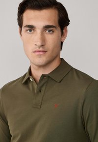 Mladý muž s krátkymi tmavými vlasmi, oblečený v olivovozelenom polo tričku s malou červenou vyšívanou nášivkou na hrudi.