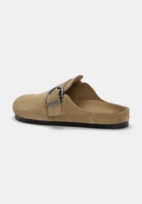 Barna velúr slip-on cipő kerek orral, ezüst csattal és fekete gumi talppal, amely texturált mintázattal rendelkezik.