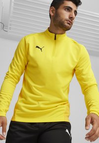 Maglietta a maniche lunghe gialla in materiale sintetico con mezzo zip, caratterizzata da un logo Puma nero sul petto e pannelli laterali in rete per la ventilazione.