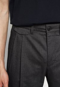 Närbild av en person som bär mörka strukturerade byxor med knäppbart midjeband och ficka med lock, i kombination med en svart skjorta.