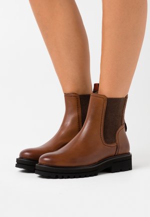 Piernas con botas Chelsea de cuero marrón con paneles laterales elásticos negros y suelas negras gruesas, de pie sobre una superficie blanca.