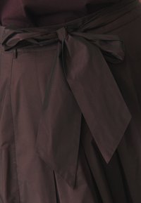 Dunkelbrauner, gefalteter Rock mit einer gebundenen Schleifen-Gürtel. Hergestellt aus glattem Stoff, mit klaren Linien und einer strukturierten Oberfläche.