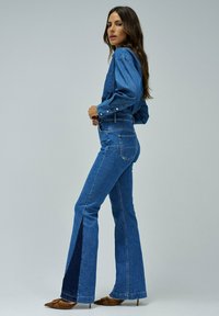 Jeans de mezclilla azul acampanados con una cintura alta, con un detalle de abertura en las partes inferiores y un material texturizado. Combinados con una camisa azul a juego.