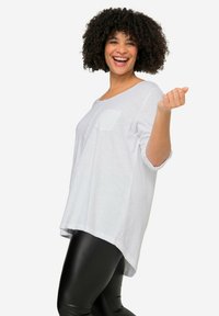 Maglietta bianca a maniche lunghe con orlo arrotondato e taschino sul petto, abbinata a leggings neri lucidi. Il modello posa con un'espressione allegra.