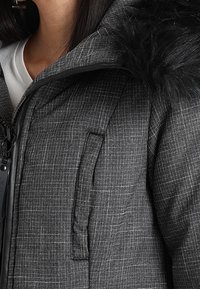 Svart pufferjacka med en texturerad grå tyg, som har en fuskpälshuva, sidoficka och dragkedjedetaljer.