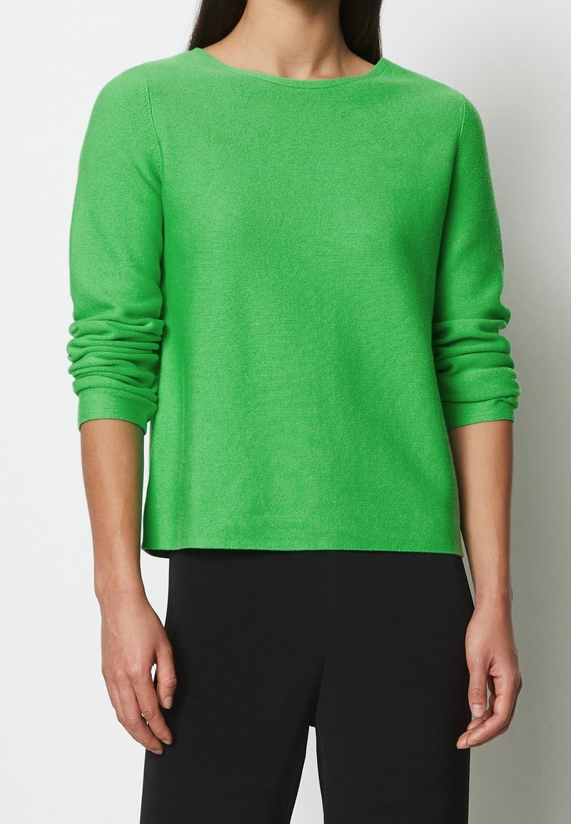 Femme portant un pull vert vif à manches longues avec des manches froncées et un pantalon noir, debout devant un fond blanc uni.