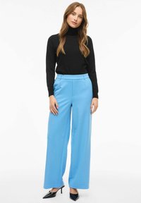 Sort rullekrave sweater kombineret med lyseblå bukser med vide ben. Bukserne har en glat tekstur og en skræddersyet pasform. Sorte højhælede sko.