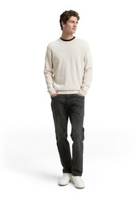 Pull beige avec des poignets côtelés, col noir ; jeans foncés, baskets blanches ; pose debout, fond neutre.