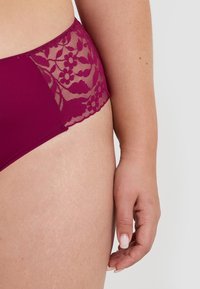 Femme portant de la lingerie en dentelle violette se concentrant sur la hanche et la main contre un fond uni.