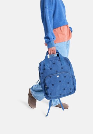 Persoon draagt blauwe trui, oranje shirt, lichtblauwe jeans en bruine schoenen, met een blauwe rugzak met zwarte bloemborduursels in de hand.