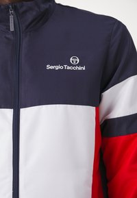 Navy, red, and white color-blocked jacket made of lightweight fabric. Features a full zip, high collar, and logo on the chest.