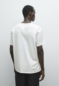 Hvid bomulds T-shirt med en afslappet pasform, rund halsudskæring og korte ærmer, båret af en model set fra ryggen mod en lys baggrund.