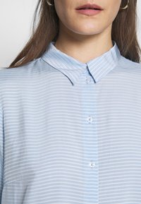 Camicia button-up azzurro chiaro con sottili righe bianche, colletto a punta e texture liscia. Presenta bottoni frontali e una leggera lucentezza.