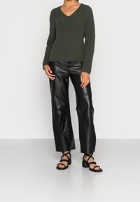 Nainen, joka pukeutuu tummanvihreään neulepuseroon, mustiin leveälahkeisiin tekonahkahousuihin ja mustiin, hihnallisiin blokki-korkokenkiin, seisoo vaalealla lattialla.