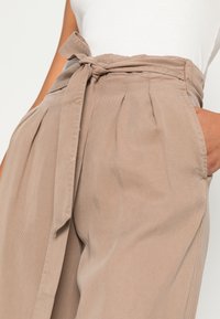 Calças bejes de cintura alta feitas de um tecido texturizado, com um cinto amarrado, pregas e bolsos laterais para um visual estruturado.
