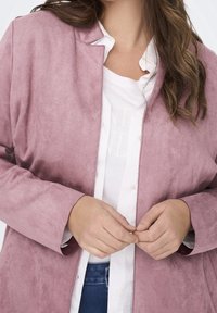 Pudrově růžová bunda z faux suede s bez límce a dlouhými rukávy, vrstvená přes bílou blůzu s knoflíkovými detaily.