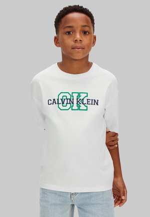 Chlapec s krátkými kudrnatými vlasy, oblečený v bílém tričku Calvin Klein s nápisy v zelené a námořnické modři, stojící na jednobarevném šedém pozadí.