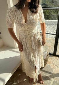 Femme portant une robe crème en dentelle boutonnée, tenant un livre, debout pieds nus sur un sol en pierre près d'une fenêtre ensoleillée avec une vue pittoresque sur l'extérieur.