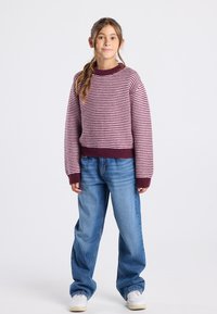 Raitapaitainen neulepusero tummanpunaisena ja vaaleanpunaisena, pyöreällä kauluksella, yhdistettynä sinisiin farkkuihin ja valkoisiin lenkkareihin. Rennosti istuva ja ribatut hihansuut.