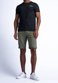 Mustikka puuvillapaita logolla, yhdistetty oliivinvihreisiin käärittyihin shortseihin ja valkoisiin nauhallisiin tennareihin, joissa on kontrastiväri pohjassa.