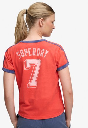 Lyshåret kvinde med hestehale iført en koralrød T-shirt med blå kant og hvid teksten "SUPERDRY 7" i stor skrift på ryggen.