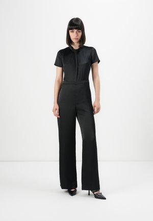 Woman with straight black hair wearing a black short-sleeve jumpsuit and black pointed-toe heels standing against white background.