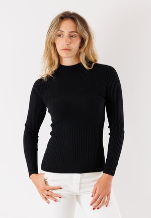 Maglione nero a coste con maniche lunghe, collo alto, silhouette aderente e dettagli testurizzati sul petto, abbinato a pantaloni bianchi.