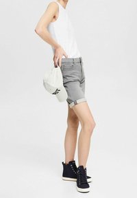 Osoba w zgiętych szarych dżinsowych szortach, białej bezrękawnik, czarnych trampkach na wysokiej podeszwie, trzymająca białą czapkę za plecami.