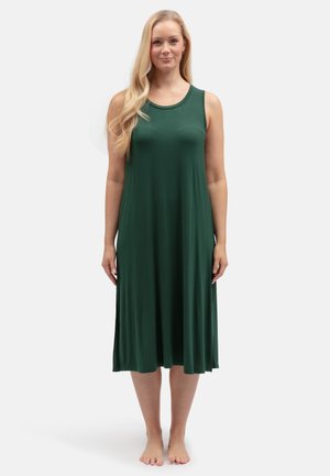 Frau mit langen blonden Haaren, barfuß stehend, trägt ein ärmelloses dunkelgrünes Midikleid vor einem weißen Hintergrund.