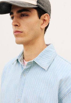 Ung mand iført en sort og beige kasket og en lyseblå denimjakke med nålestriber, der ser eftertænksomt mod venstre.