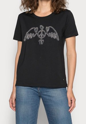 Woman wearing black t-shirt with shiny silver peace symbol and wings design, paired with blue jeans.