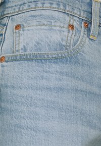 Poche de jean en denim bleu clair avec couture jaune visible, rivets en cuivre et détail de passants de ceinture.