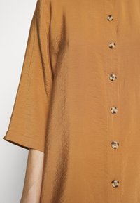 Robe marron à manches courtes avec une texture froissée, ornée d'une rangée de cinq grands boutons ronds sur le devant.