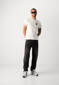 Lyscremet bomulds t-shirt med et farverigt grafisk design på brystet, parret med mørke denim jeans og grå sportsko.