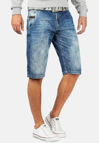 Shorts en jean bleu délavé avec une coupe slim, dotés de poches avant et d'une poche zippée. Ourlet replié et couture visible. Associés à des baskets blanches.