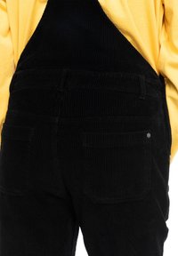 Schwarze Corduroy-Latzhose mit sichtbaren Nähten und zwei Gesäßtaschen, kombiniert mit einem gelben Langarmhemd. Die Textur ist gerippt und kuschelig.