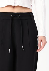 Pantaloni neri con vita regolabile tramite coulisse e punte in metallo argentato, design plissettato e texture liscia, abbinati a un top bianco cropped.