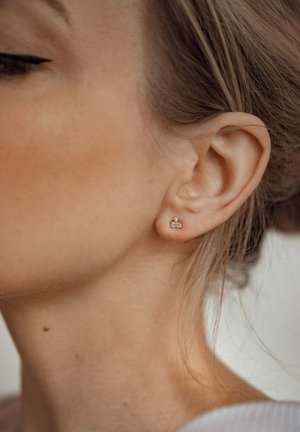Złota kolczyk wkrętka z małym prostokątnym przezroczystym kamieniem, noszona na uchu, prezentująca minimalistyczny design na tle jasnej karnacji.
