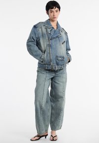 Een persoon staat zelfverzekerd, gekleed in een grote spijkerjack en losse jeans, in combinatie met zwarte slippers, tegen een effen achtergrond.