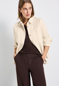Beżowa koszula-kurtka na guziki z dwiema kieszeniami na piersi, z podwiniętymi rękawami, w połączeniu z ciemnobrązową bluzką i dopasowanymi spodniami. Gładka tekstura.