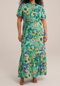 Maxiklänning i grönt med blommönster i gult och lila, korta ärmar, ett bälte vid midjan och en lager-på-lager fåll. Tillverkad av lättviktig tyg.