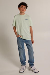 T-shirt in cotone verde chiaro con maniche corte, con testo blu. Indossato con jeans blu a vestibilità sciatta e sneaker bianche.