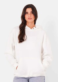 Vrouw met lang donker haar, gekleed in een wit sweatshirt met capuchon en lichtblauwe spijkerbroek, handen in de zakken van het sweatshirt, neutrale achtergrond.