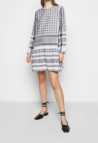 Femme portant une robe à manches longues à motifs noir et blanc, associée à des chaussures plates à lacets noires, se tenant contre un fond clair et uni.